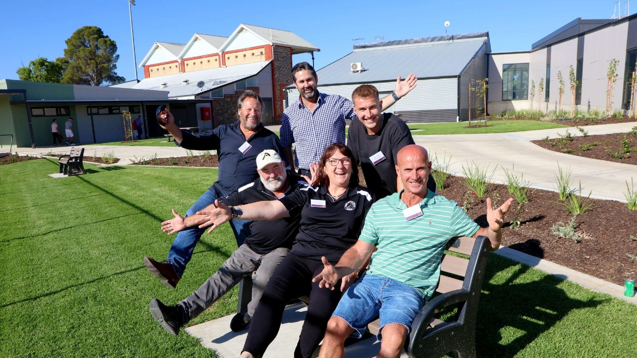 Group of people smiling with excitement sitting on park bench on lovely sunny day with green grass around them and new park buildings behind them.