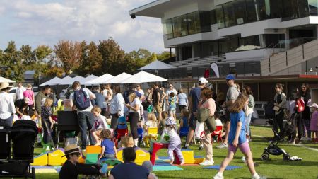 Crowd of people enjoying socialising and picnics on green grass on large oval on bright sunny day. New oval seating and grandstand are up to their right.