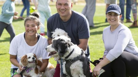 Three people kneeling and smiling with their dogs in front of them on green grassy oval on sunny day. Children are playing in the background.