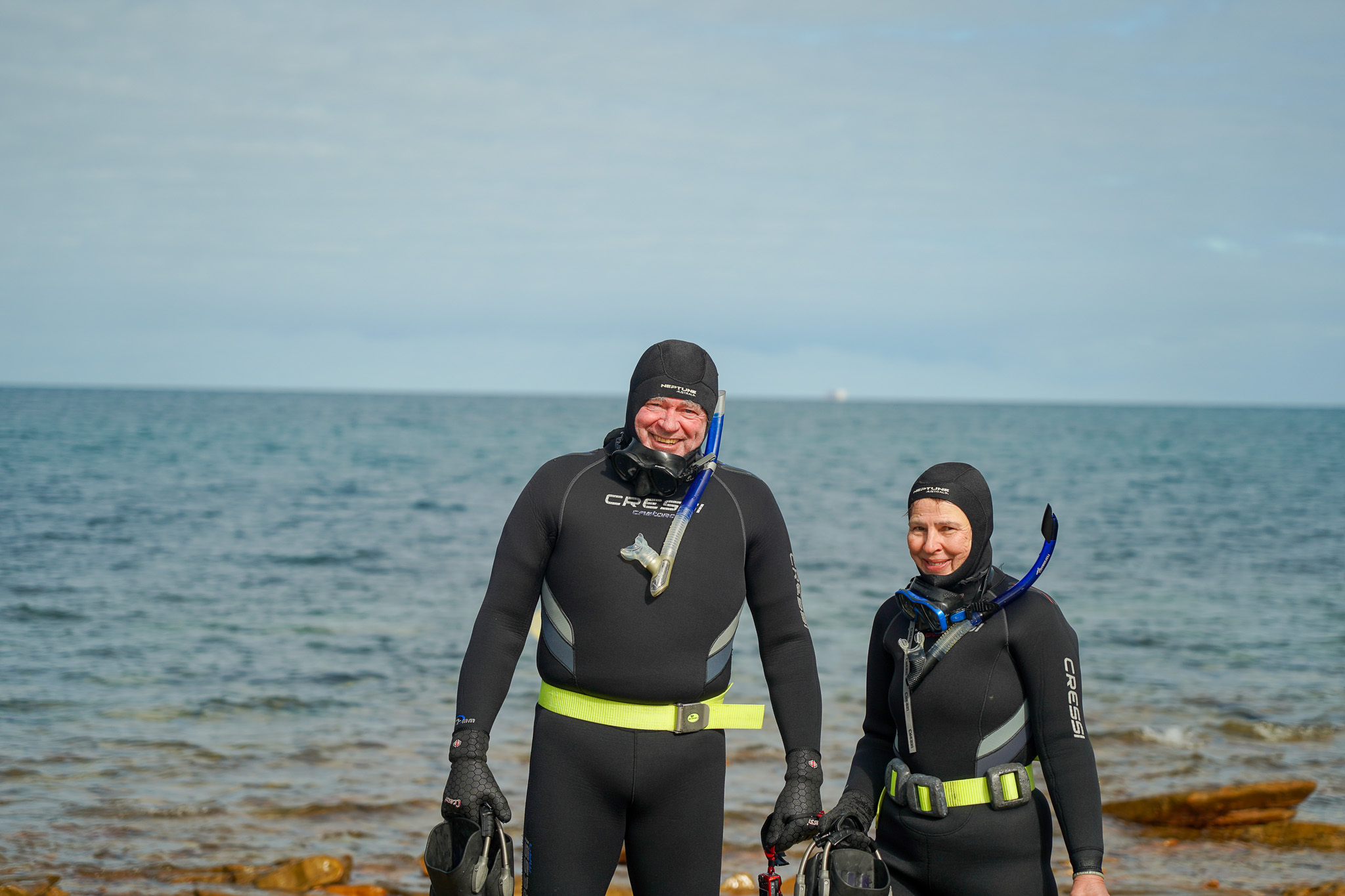 2 people in wetsuits and snorkelling gear standing by the ocean
